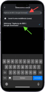 Captura de tela da interface de seleção de local em um aplicativo móvel, exibida em português. No topo, há uma barra de busca com o texto "Agência de SEO | Google Dominado" digitado. Abaixo, aparece a indicação "Local é uma residência (casa)", seguida por uma sugestão destacada com uma seta verde: "Adicionar 'Agência de SEO | Google Dominado'", acompanhada de um ícone de marca de seleção azul, indicando que a opção foi selecionada. O teclado virtual está visível na parte inferior da tela, sugerindo que o usuário está em processo de confirmação ou edição do nome do local. A imagem representa a etapa de inclusão ou validação de um ponto comercial em uma plataforma de localização ou cadastro empresarial.